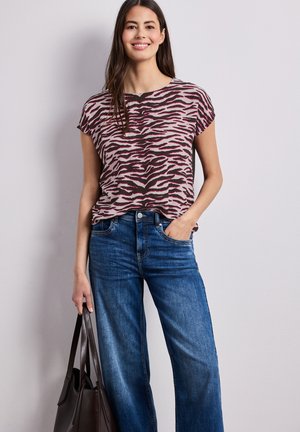 Kvinde smiler iført en pink og sort zebrastribet kortærmet top, blå jeans og holder en mørkebrun lædernettaske.