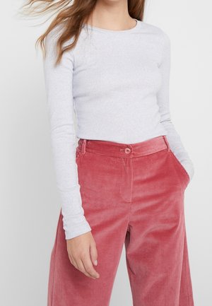 Vrouw die een nauwsluitend lichtgrijs shirt met lange mouwen draagt, ingestopt in een hoge taille, wijd uitlopende rode corduroy broek, met haar hand in de zak.