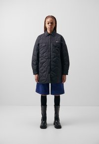 Black quilted jacket with a button front, two chest pockets, and a small logo. Paired with denim shorts, black leggings, and black ankle boots.