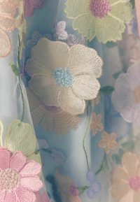 Ljusblått tyg med intrikata blomsterbroderier i pastellfärger, med detaljerade kronblad och stjälkar som skapar en texturerad design.