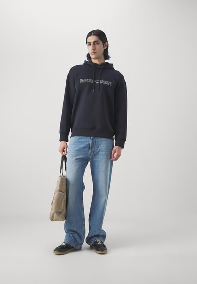 Felpa navy con logo "Emporio Armani", jeans flared blu chiaro, sneakers nere e borsa beige con tracolla. Texture morbide.