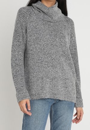 Femme portant un pull à col roulé en maille grise et un jean bleu, debout devant un fond clair uni.