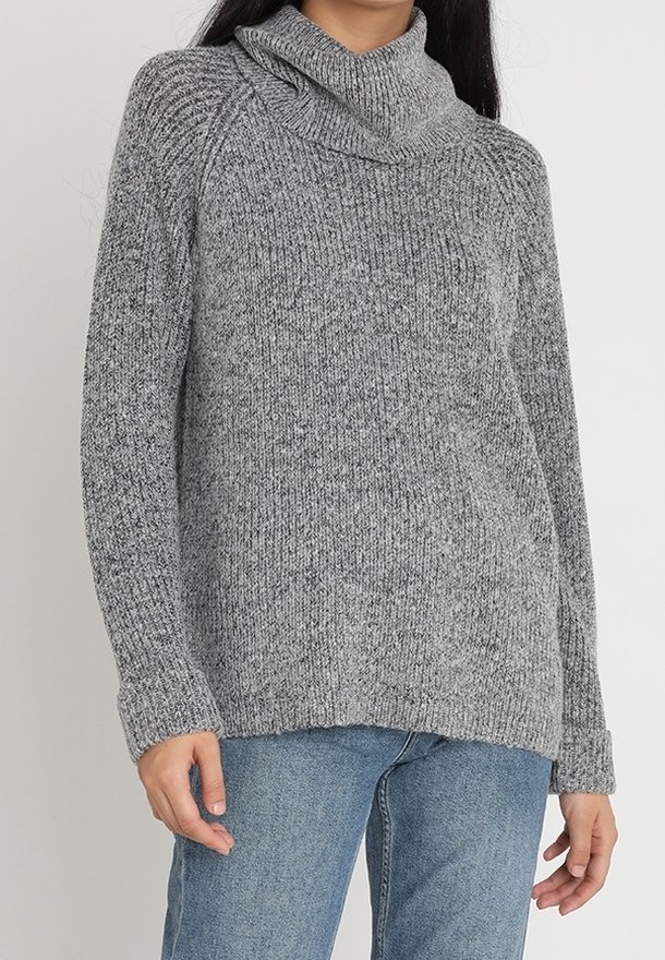 Femme portant un pull à col roulé en maille grise et un jean bleu, debout devant un fond clair uni.
