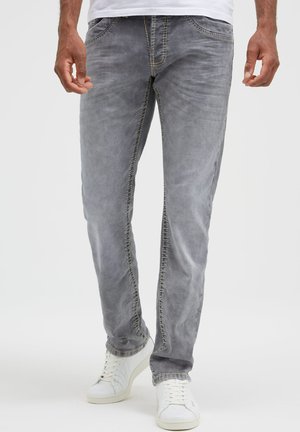 Mann trägt ausgewaschene graue Jeans und weiße Sneaker, steht mit locker neben dem Körper hängenden Händen vor einem einfarbigen Hintergrund.