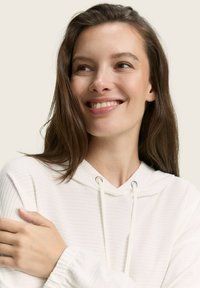 Junge Frau mit langen braunen Haaren, die lächelt und zur Seite schaut, trägt einen weißen gerippten Hoodie mit Kordelzügen.