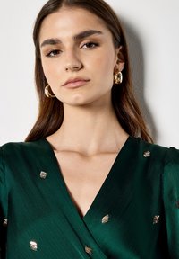 Groene wikkeljurk met een gladde textuur, versierd met gouden pailletten. Haar stijl recht, met ronde gouden oorbellen.