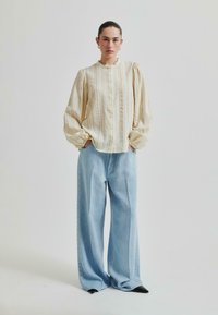 Svetlo obarvana, prugasta bluzica s gumbom z mehkim občutkom in napihnjenimi rokavi, v kombinaciji s širokimi svetlo modrimi jeans hlačami in črnimi koničastimi čevlji.