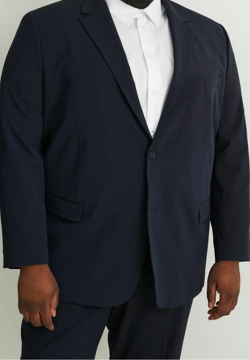Homme portant une veste de costume bleu marine foncé et un pantalon assorti avec une chemise blanche boutonnée jusqu'en haut, sur un fond uni.