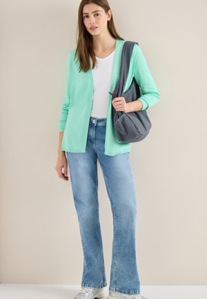 Mujer con cárdigan verde menta sobre camisa blanca, vaqueros azules, zapatillas plateadas, sosteniendo un bolso gris al hombro sobre fondo beige.