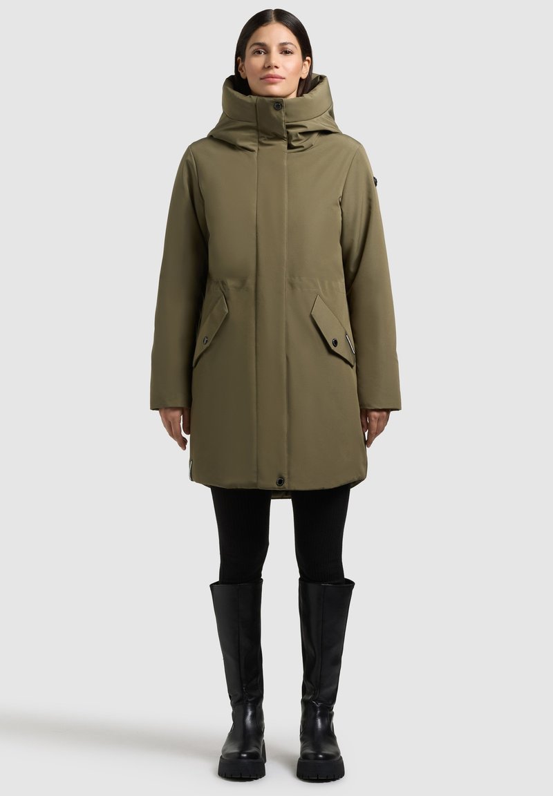 Oliwkowo-zielony ocieplany płaszcz z wysokim kołnierzem, dwoma kieszeniami z patkami z przodu oraz suwakiem na przodzie. Noszony na czarnych legginsach i kozakach za kolano.