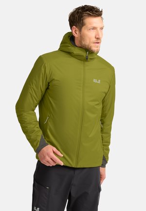 Man draagt een groene outdoorjas met capuchon en ritssluitingen aan de zakken en zwarte broek, kijkt naar rechts.
