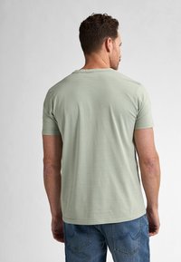 Camiseta de algodón verde claro con mangas cortas y escote redondeado, vista desde atrás, combinada con unos pantalones vaqueros azules.