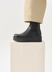 Botină neagră din piele, cu talpă groasă neagră, având panouri laterale elastice, purtată cu pantaloni bej, poziționată pe un suport deschis.