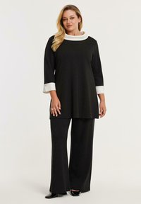 Frau trägt eine schwarze Langarm-Tunika mit weißem Kragen und Manschetten, kombiniert mit einer schwarzen weiter Hose und schwarzen spitzen Schuhen.