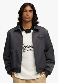 Giubbotto bomber grigio con chiusura a zip, polsini a coste e la parola "Represent" ricamata sul petto, indossato sopra una camicia bianca.