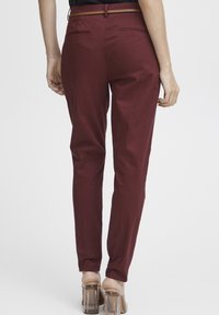 Pantalons ajustés bordeaux avec une texture lisse, dotés de poches arrière et d'une ceinture marron assortie. Des plis nets au niveau de la cheville soulignent la silhouette.