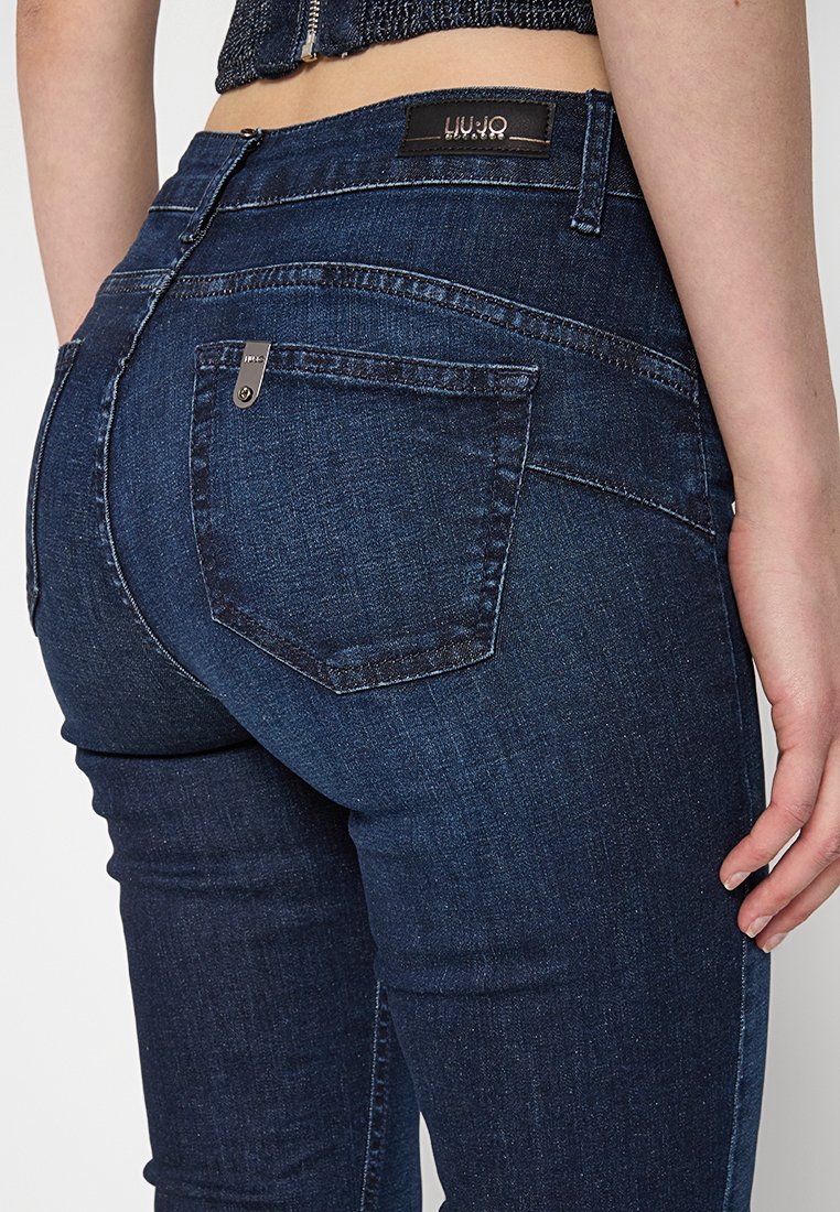 Jeans skinny in denim blu scuro con una texture liscia, dotati di una tasca posteriore, accessori metallici e un'etichetta con logo in vita.