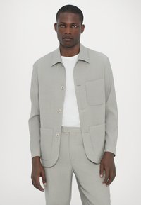 Gaiši pelēks žakets ar apkakles kakla izgriezumu, priekšējiem pogām, divām plecu kabatām, kas kombinēts ar baltu kreklu un atbilstošām biksēm. Gluda tekstūra.