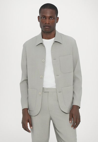 Ανοιχτό γκρίζο σακάκι με γιακά, μπροστινά κουμπιά, δύο τσέπες με πατίνι, συνδυασμένο με λευκό πουκάμισο και αντίστοιχο παντελόνι. Λείος υφασμάτινος σχεδιασμός.
