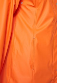 Veste imperméable orange en matière lisse et brillante, avec boutons pression et une couture continue sur le côté.