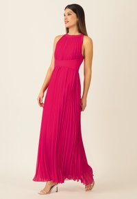 Langes, plissiertes Abendkleid in leuchtendem Pink mit hohem Kragen und breitem Taillenbund. Der fließende Stoff erzeugt eine sanfte, strukturierte Silhouette.