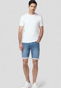 Biała bawełniana koszulka, jasnoniebieskie szorty dżinsowe z podwiniętymi nogawkami i białe sneakersy. Model stoi na neutralnym szarym tle.