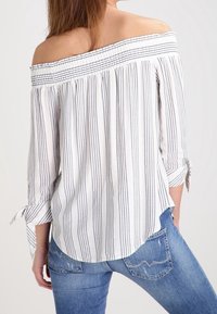 Off-shoulder vit topp med blå vertikala ränder, med smockad halsringning och trekvartslånga ärmar med knytning, tillsammans med blå jeans.