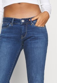 Los jeans de mezclilla azul tienen una forma ajustada, un diseño de cinco bolsillos y costuras amarillas en contraste en las costuras y la cintura.