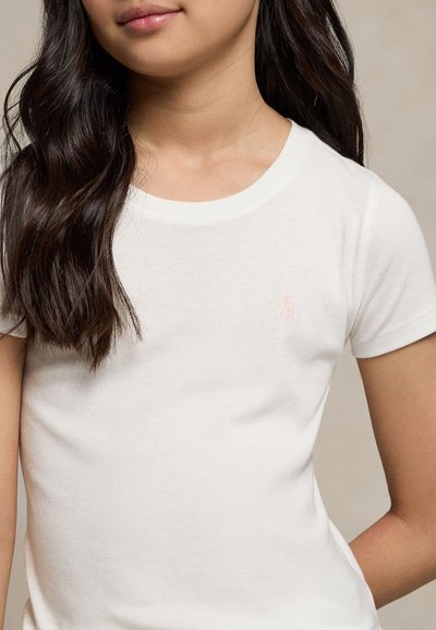 Jeune personne aux longs cheveux foncés portant un t-shirt blanc uni à manches courtes avec un petit logo brodé sur la poitrine.