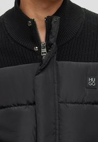 Giubbotto imbottito nero con colletto alto a coste, chiusura con zip e patch logo tono su tono. Il tessuto presenta una superficie liscia e un design trapuntato.