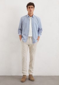 Hellblau gestreiftes Button-Up-Hemd mit einem weißen Unterhemd, beigen Hosen und braunen Wildlederschuhen. Modell posiert mit Händen in den Taschen.