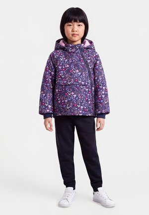 Enfant debout portant une veste à capuche fleurie violette, un pantalon noir et des baskets blanches sur un fond clair uni.
