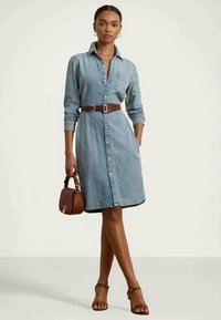 Denimskjortekjole med lange ermer, knappelukking foran og brystlomme, innsnevret i midjen med et brunt belte. Brun håndveske og sandaler.