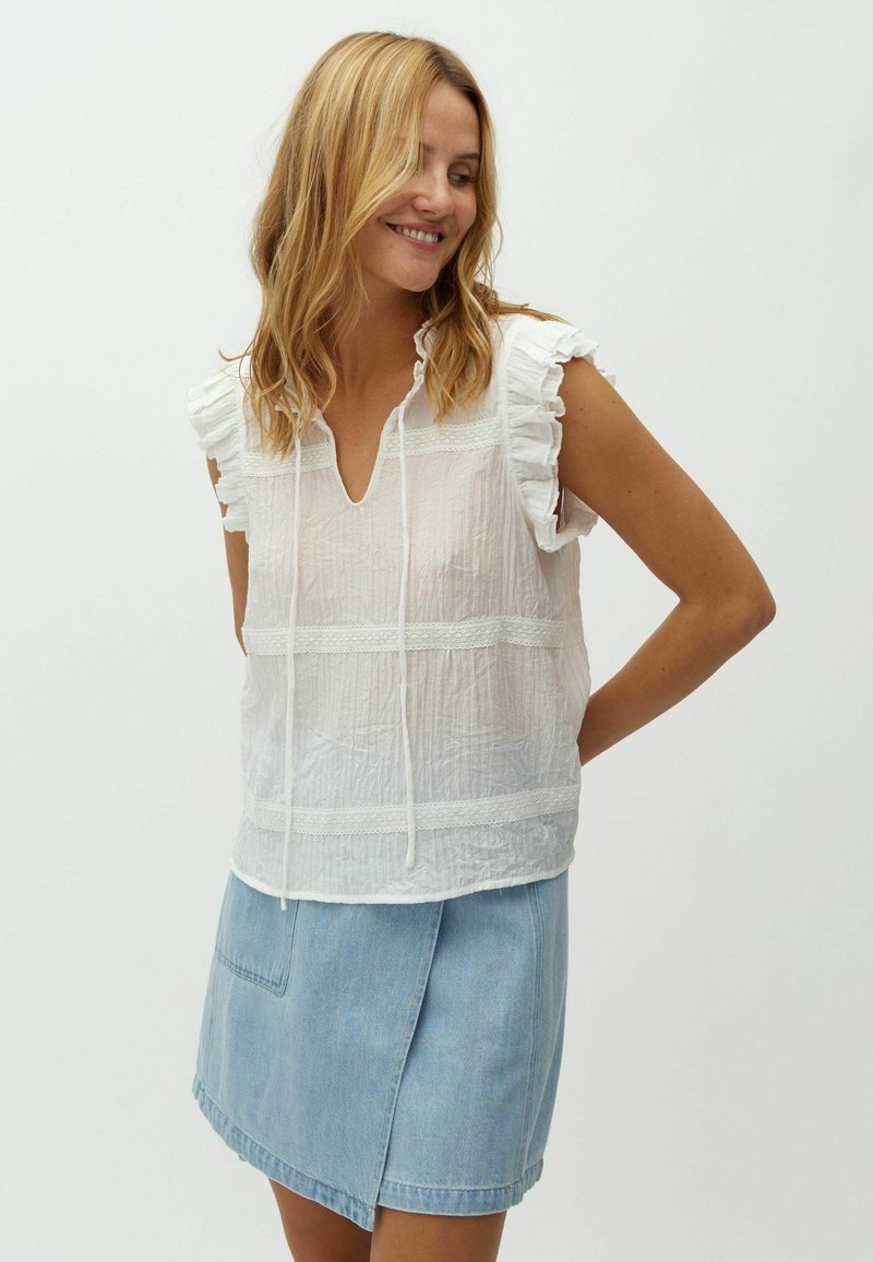 Femme souriante aux cheveux blonds portant un chemisier blanc sans manches avec des volants et une jupe en jean bleu clair, debout devant un fond uni.