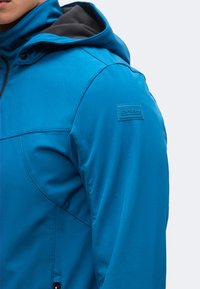 Veste softshell bleue avec capuche noire, coutures visibles et petite étiquette rectangulaire de la marque sur la partie supérieure de la manche.