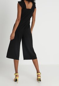 Svart jumpsuit med korta ärmar, midikjol, strukturerad tygkvalitet och dragkedja i ryggen. Ett par gula klacksandaler fulländar looken.