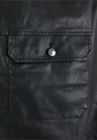 Detail eines schwarzen Lederjackets mit einer aufgesetzten Tasche und einem silbernen Druckknopf, glatter Textur und sichtbaren Nähakzenten.