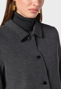 Dark grey sweater with a collared neckline, featuring a textured knit and three black buttons. Worn over a dark turtleneck. Gold earrings visible.