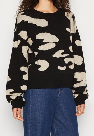 Femme portant un pull noir avec un motif abstrait camouflé beige et un jean en denim bleu, debout devant un fond uni.
