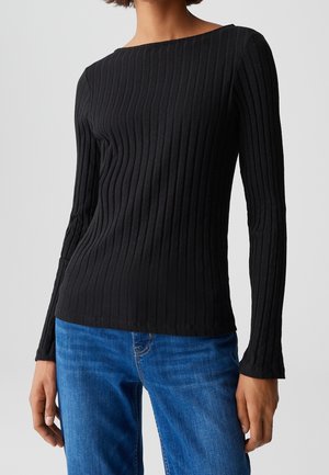 Donna che indossa una maglia nera a coste a maniche lunghe abbinata a jeans blu, in piedi davanti a uno sfondo chiaro semplice.