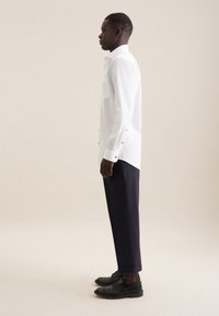 Mand stående i profil iført en hvid langærmet skjorte, mørke korte bukser og sorte pæne sko på en ensfarvet baggrund.