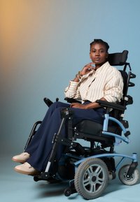 Fauteuil roulant électrique avec un siège noir, un cadre bleu et des roues grises. L'utilisateur porte une chemise rayée et un pantalon bleu marine, posé avec confiance dans un studio.
