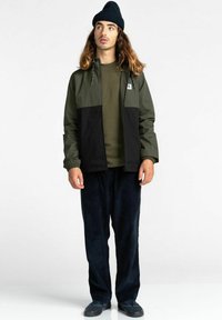 Mann in einer dunkelgrünen und schwarzen Jacke, olivgrünem Hemd, schwarzer Mütze, lockeren schwarzen Hosen und schwarzen Sneakern, der steht und seitlich schaut.