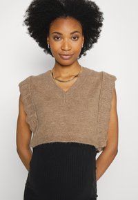 Suster sem mangas em tricô castanho com bordas em escamas, design em V e uma textura suave, usado sobre uma saia preta de canelados.