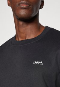 Svart sweatshirt med strukturerad tyg, med en liten vit logotyp "JJ90" och triangelaccent på bröstet. Avslappnad passform, rundhalsad design.