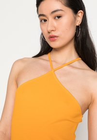 Top amarelo sem mangas com uma textura canelada, apresentando alças assimétricas. O modelo usa brincos de argola e tem cabelo liso e escuro.