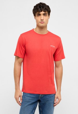 Man with dark curly hair wearing a red Mustangs t-shirt and blue jeans, standing against a plain light background.