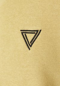 Żółta tkanina o gładkiej fakturze z czarnym haftowanym logo w kształcie trójkąta w prawym dolnym rogu.