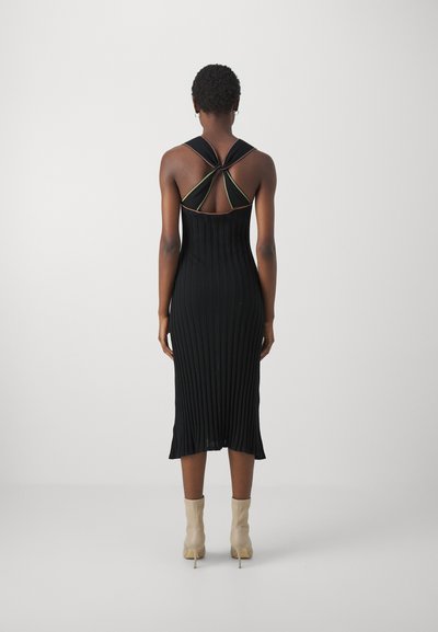Robe midi noire à côtes avec une silhouette ajustée, dos ouvert avec des bretelles croisées et une texture subtile. Portée avec des bottines beiges.
