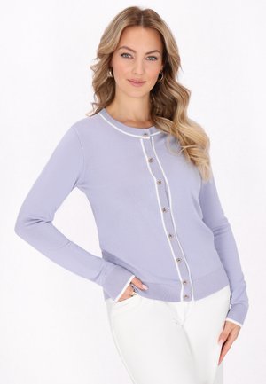 Femeie cu păr lung, ondulat, blond, purtând un cardigan mov deschis cu nasturi, cu margini albe și pantaloni albi, stând cu o mână în buzunar.